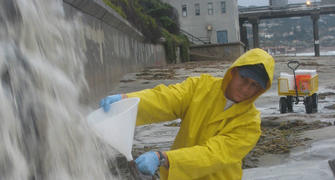 A researcher in a yellow raincoat uses a funnel to collect water from storm water runoff at a beach.