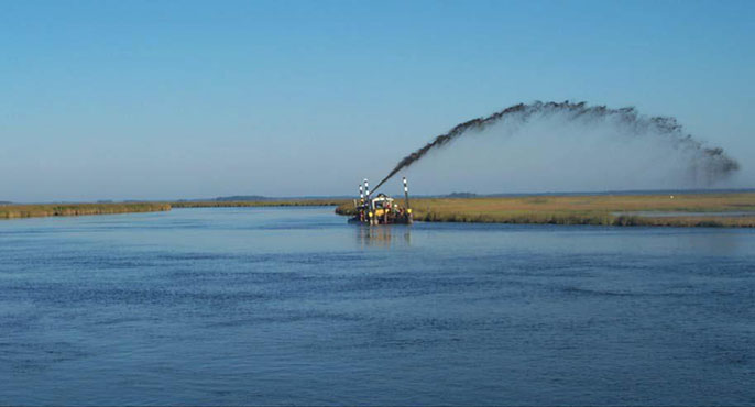 Sediment is shot out of a machine in a wide arc across the sky, a technique that enables the sediment to be spread across a low-lying coastal wetland area as part of a pilot effort to raise its elevation.