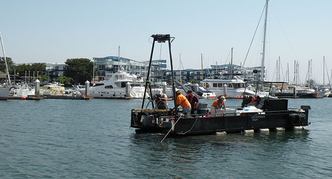 A five-member field crew travels through Marina del Rey Harbor on a research vessel to collect sediment grab samples from the harbor’s seafloor. In the background are docked boats and yachts.