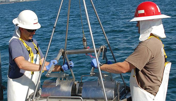 Two field researchers standing at the back of a research vessel in San Diego Bay lower a sediment grab sampler to the seafloor to collect sediment samples.