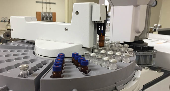 A circular tray holds vials containing water samples that will be analyzed via a chromatography-mass spectrometry instrument, which is visible in the background. 