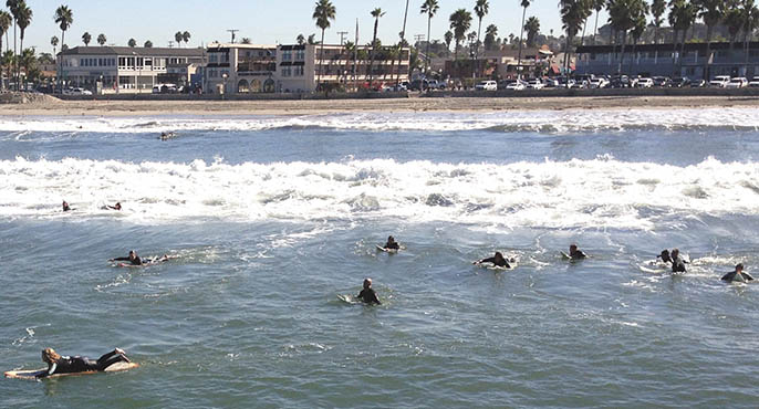Surfers laying on their surfboards paddle away from the shoreline at Ocean Beach in San Diego, with homes and businesses along the shoreline visible in the background. 