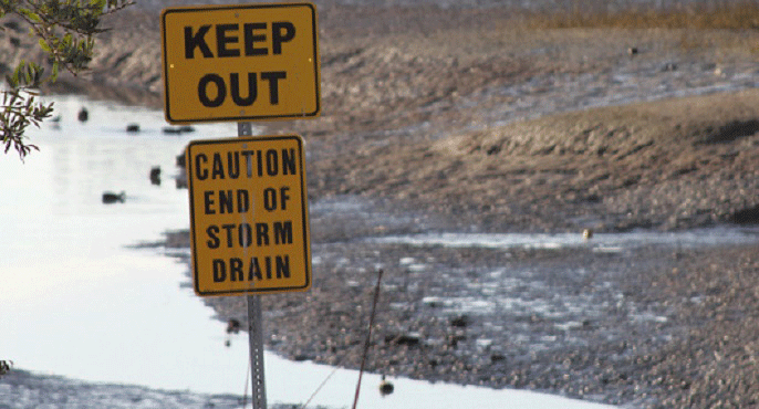 At the terminus of a storm drain in Newport Bay in Orange County, a pair of yellow signs reads: "Keep out" and "Caution -- End of Storm Drain." 