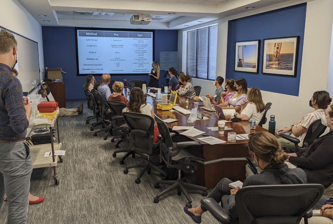 A group of about 20 researchers listens to a presentation about environmental DNA in a SCCWRP conference room with a large table in the middle.