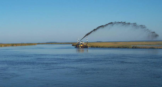  Sediment is shot out of a machine in a wide arc across the sky, a technique that enables the sediment to be spread across a low-lying coastal wetland area as part of a pilot effort to raise its elevation. 